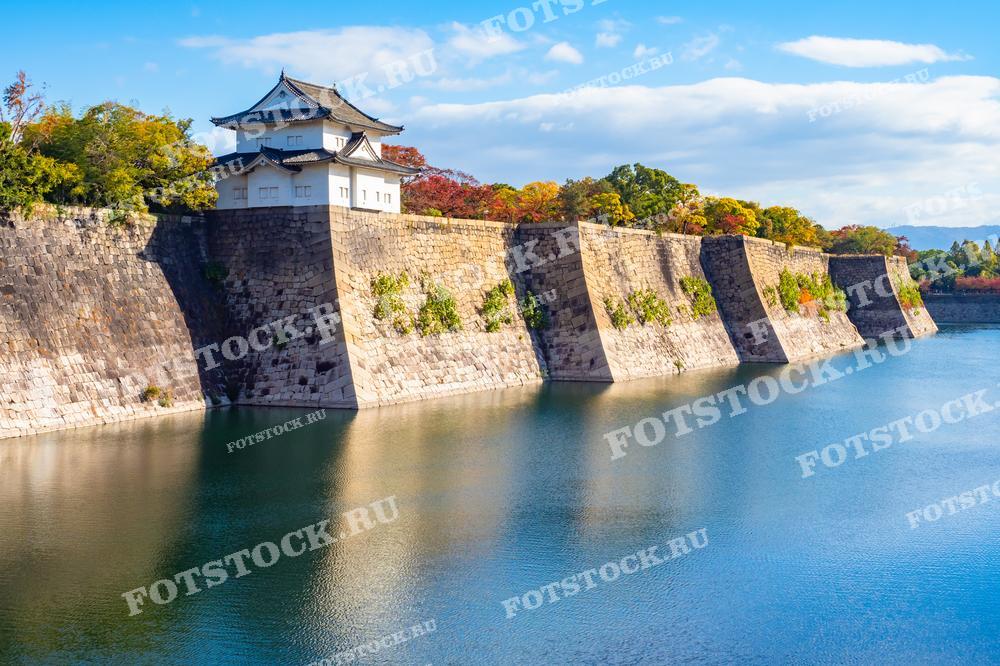 На фотографии изображена крепостная стена Осакского замка с прилегающим водным рвом. Осенние деревья на заднем плане придают яркие краски древним каменным стенам, создавая контраст с отражением в воде. Яркий солнечный свет подчеркивает детали архитектуры и природного окружения.