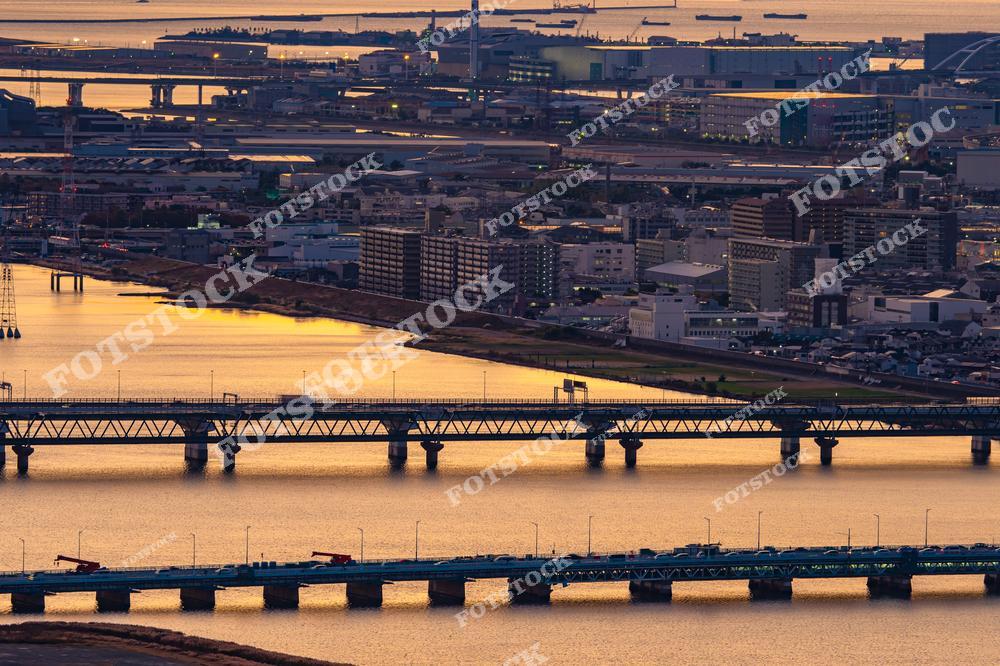 На фотографии изображены мосты, пересекающие реку в Осаке, освещённые мягким закатным светом. Отражения на водной глади и индустриальный фон подчёркивают красоту и масштаб городской инфраструктуры. Осака, один из крупнейших портовых городов Японии, славится своими мостами и развитой транспортной системой. Закатное освещение добавляет сцене тёплые оттенки, создавая романтичное и спокойное настроение. Этот вид символизирует гармонию между природой и индустриальным ландшафтом.