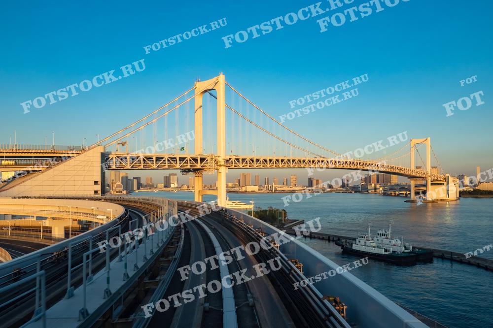 Фотография запечатлела Радужный мост в Токио во время заката. Этот подвесной мост соединяет районы города через Токийский залив, являясь важной частью транспортной инфраструктуры. Вид на мост и железнодорожные пути в сочетании с закатным светом создаёт впечатляющий городской пейзаж.