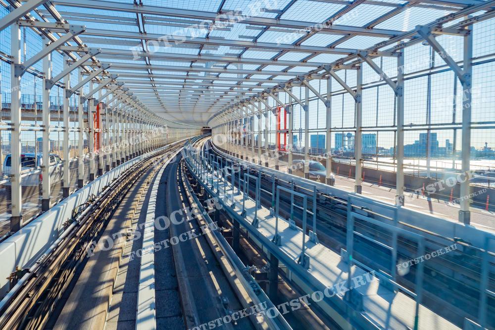 Фотография демонстрирует изогнутую трассу монорельса на мосту в Токио. Современная конструкция из стекла и металла создаёт тоннель, по которому движется поезд, окружённый городскими пейзажами. Светлое дневное освещение подчёркивает детали инфраструктуры.