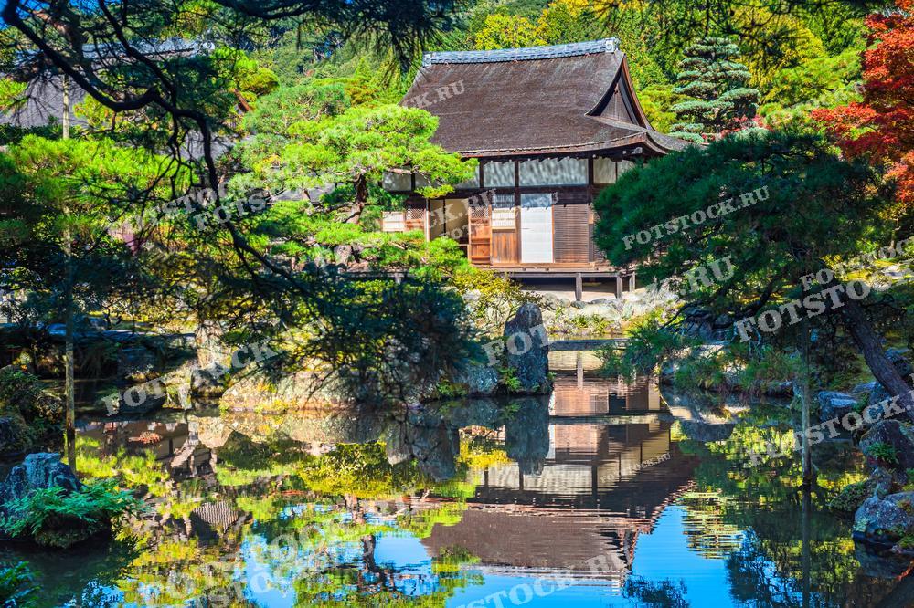Фотография изображает храм Гинкаку-дзи в Киото, также известный как Серебряный павильон. Вокруг храма расположен традиционный японский сад с деревьями и прудом, в котором отражаются здание и природа. Этот храм известен своей спокойной атмосферой и гармонией с окружающей средой.
