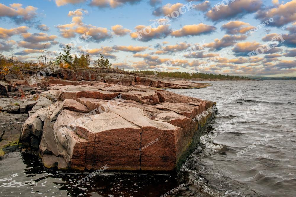 На фотографии запечатлён живописный берег Ладожского озера, известный своими гранитными выходами. Красные и серые скалы создают уникальный ландшафт, характерный для карельских шхер. Осенние деревья и облачное небо подчёркивают суровую красоту природы Карелии. Волны неспешно накатывают на каменные берега, создавая атмосферу спокойствия и уединённости.