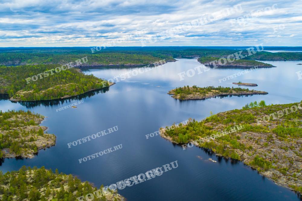 На фотографии изображен вид с дрона на Ладожские шхеры в Карелии. Острова, покрытые густым лесом, плавно уходят в воду Ладожского озера. Спокойная водная поверхность отражает облачное небо. Карелия известна своими живописными природными ландшафтами и является популярным местом для туристов и любителей природы.