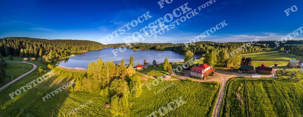 На фотографии изображён сельский пейзаж Карелии с высоты. В центре кадра находится небольшое озеро, окружённое зелёными полями и хвойными лесами. Деревенские дома и хозяйственные постройки создают атмосферу уюта и спокойствия. Тёплые лучи вечернего солнца освещают природу, подчёркивая летнюю красоту карельского края. Это место идеально подходит для отдыха на природе и уединённых путешествий.