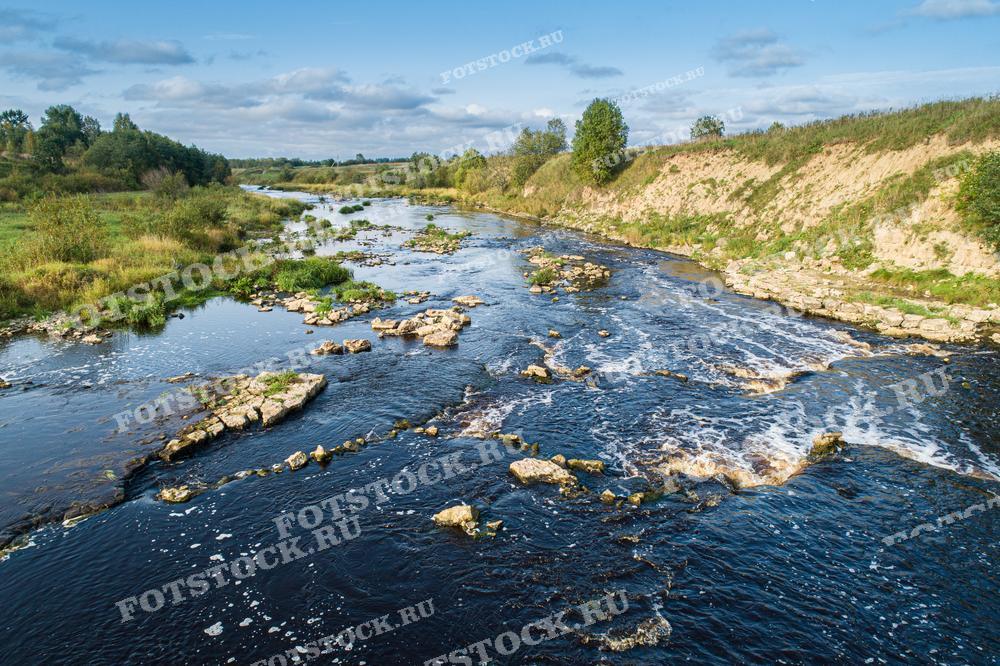 На фотографии изображена река с порогами в Карелии, окруженная зелеными берегами. Вода течет через камни, создавая небольшие водопады.
