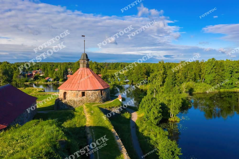 На фотографии изображена крепость Корела, расположенная в городе Приозерск. Это одно из старейших оборонительных сооружений на территории Карелии, основанное ещё в Средние века. Вид с высоты открывает панораму крепостных стен, башен и окружающей природы. Зелёные деревья и отражение голубого неба в воде подчёркивают красоту этого исторического места. Крепость Корела является популярной туристической достопримечательностью и объектом культурного наследия.