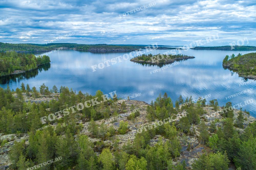На фотографии изображен вид на Ладожские шхеры, сделанный с высоты. Чистые воды Ладожского озера омывают скалистые берега и небольшие острова, покрытые густыми лесами. Ладожские шхеры — это уникальный природный объект, представляющий собой архипелаг из множества островов, которые создают неповторимые пейзажи. Это место привлекает любителей природы и фотографов со всего мира, предлагая уединение и живописные виды, особенно когда небо и вода сливаются в гармонии.