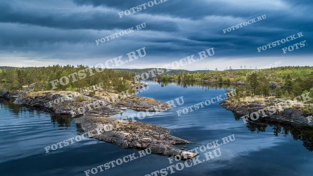 На фотографии изображены Ладожские шхеры — уникальные скалистые острова на Ладожском озере в Карелии. Вид сверху позволяет оценить красоту и суровость этого природного ландшафта. Грозовые облака создают драматичное настроение, подчеркивая дикость и нетронутость природы. Ладожское озеро — крупнейшее в Европе, а его шхеры — одно из самых живописных мест в России, привлекающее туристов со всего мира.