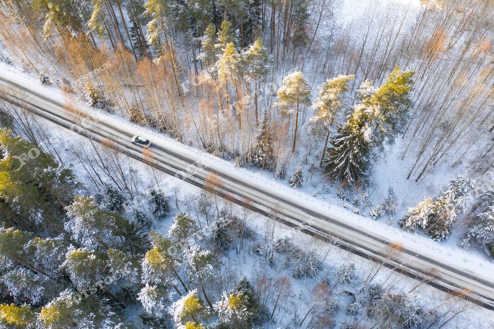 На фото изображена зимняя дорога, пролегающая через заснеженный лес в Карелии. Деревья покрыты снегом, а солнце освещает верхушки, создавая игру света и тени. Машина на дороге подчеркивает атмосферу спокойствия и уединенности северной природы. Вид сверху позволяет оценить красоту зимнего пейзажа и протяженность трассы среди лесных массивов.
