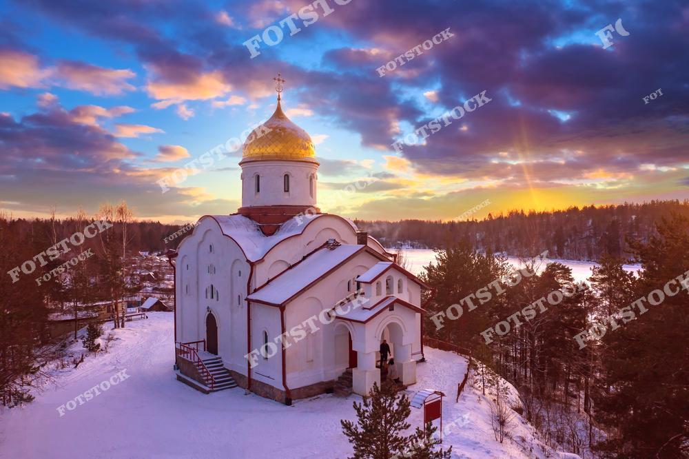 На фотографии изображена православная церковь с золотым куполом, окружённая заснеженной природой Карелии. Вид с высоты открывает живописный зимний пейзаж с лесами и замёрзшей рекой. Закатное солнце окрашивает небо в тёплые оттенки, создавая атмосферу уюта и спокойствия. Это место привлекает туристов своей красотой и духовной атмосферой.