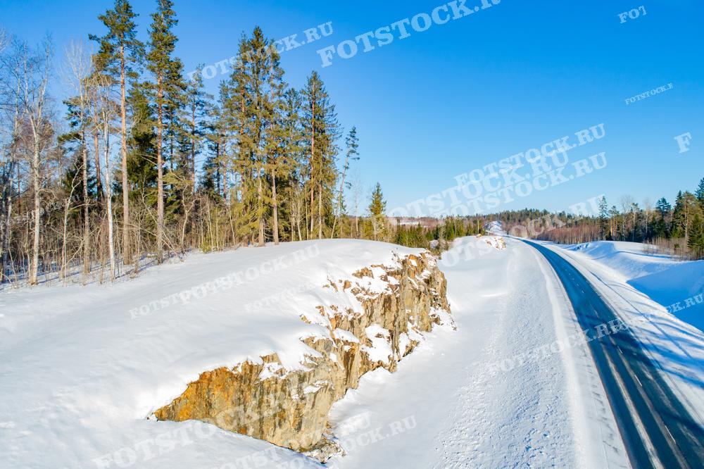 Фотография показывает дорогу в Карелии, окруженную заснеженным лесом. Скалы вдоль дороги и деревья покрыты толстым слоем снега. Ярко-голубое небо создает впечатляющий контраст с белизной снега. Эта дорога является частью живописного маршрута по Карелии, привлекающего путешественников в зимний сезон.