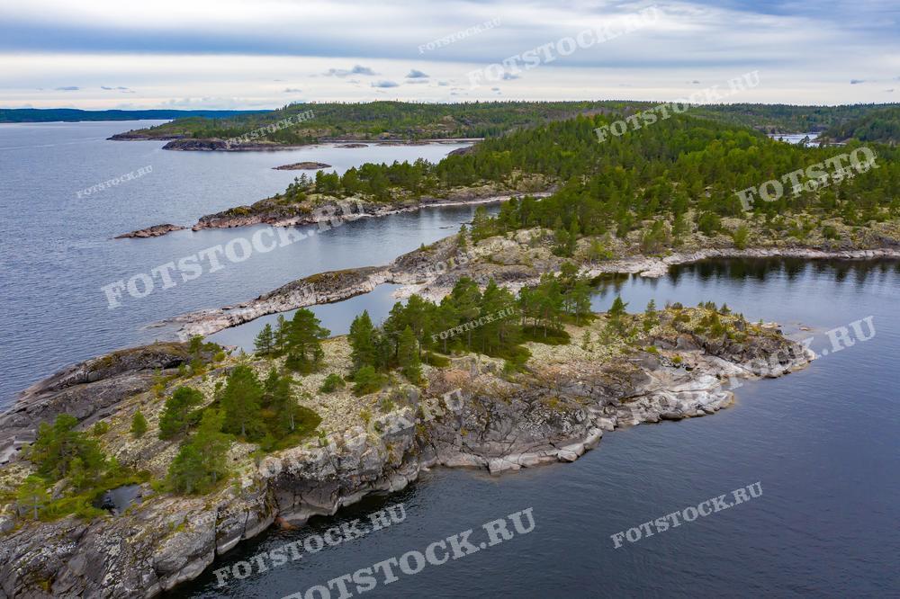 Фотография показывает вид с дрона на острова Ладожского озера в Карелии в облачный день. Каменистые берега островов покрыты густыми зелеными деревьями, создавая контраст с серым небом. Спокойная водная гладь отражает облачное небо, добавляя глубины и спокойствия в пейзаж. Ладожские шхеры – популярное место среди туристов благодаря своей уникальной природе и живописным видам.