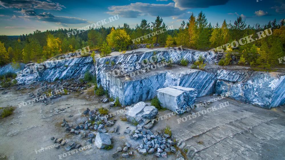 На фотографии изображен мраморный карьер в горном парке Рускеала, который когда-то был важным местом добычи мрамора. Сегодня он представляет собой популярную туристическую достопримечательность, окруженную густыми лесами, которые осенью окрашиваются в яркие цвета. Гладкие поверхности скал и разломы мрамора свидетельствуют о богатой истории этого места. Рускеальский мрамор использовался в строительстве многих знаменитых сооружений в России, включая Исаакиевский собор в Санкт-Петербурге.