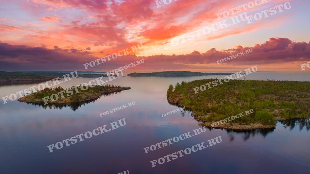 Фотография демонстрирует закат над Ладожским озером в Карелии, снятый с высоты. Острова, покрытые зелеными деревьями, окружены спокойной водой, отражающей яркие розовые и оранжевые облака. Этот живописный вид подчеркивает природную красоту Карелии и ее уникальные ландшафты, привлекающие множество туристов.