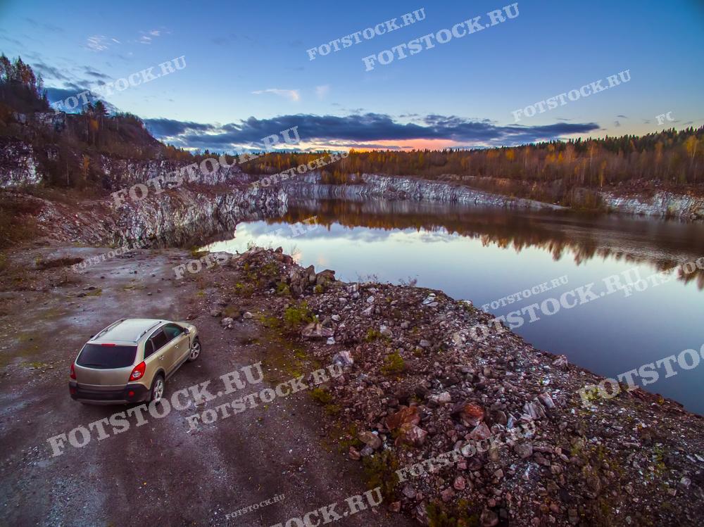 Фотография показывает Рускеалу в Карелии на фоне заката. Автомобиль припаркован на берегу, добавляя элемент современности в природный ландшафт. Спокойная вода озера отражает красочное закатное небо и облака. Карелия известна своими уникальными природными объектами, такими как этот карьер, которые привлекают туристов.