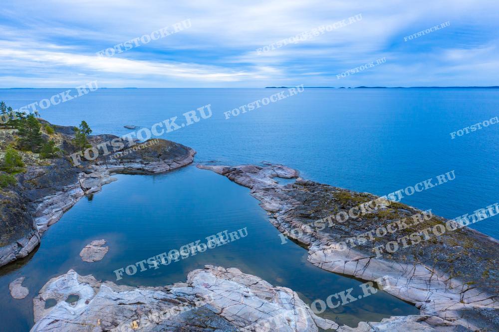 На фотографии изображен каменистый берег Ладожского озера в Карелии. Вода в озере спокойная, отражает голубое небо и облака. Снимок демонстрирует естественную красоту и тишину карельской природы.