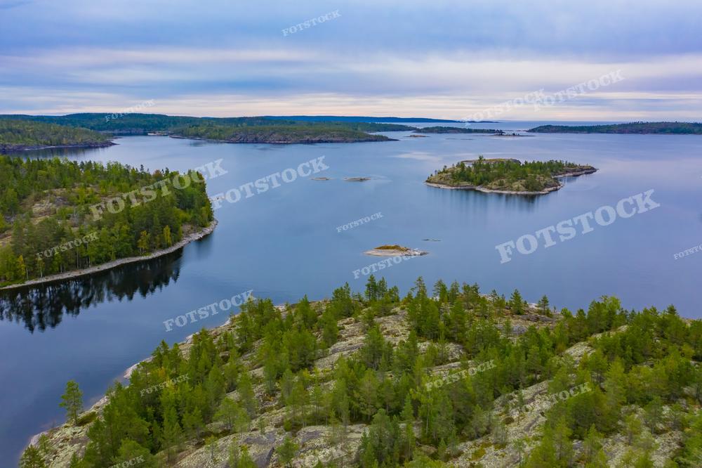 На фотографии с высоты птичьего полета запечатлены острова на фоне спокойной водной глади в Карелии. Зеленые хвойные леса покрывают острова и берега, создавая контраст с синевой воды. Небо пасмурное, что придает изображению немного меланхоличное настроение. Это место может привлекать путешественников, искателей тихих и уединенных уголков природы, а также фотографов, желающих запечатлеть уникальную красоту северных ландшафтов.