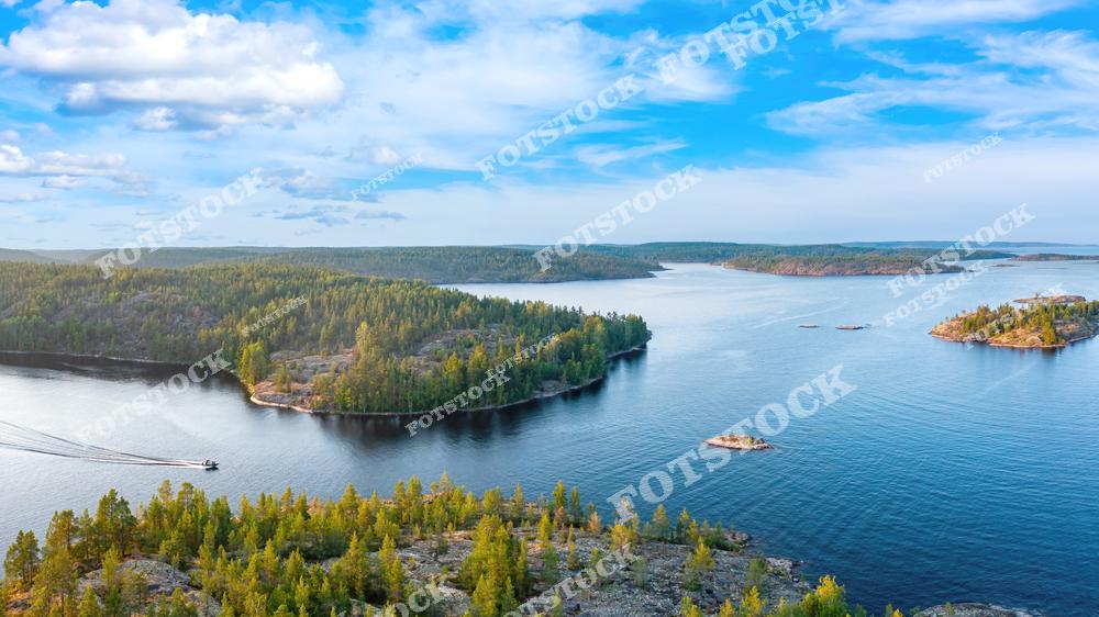 На фотографии запечатлены Ладожские шхеры в летний день, снятые с высоты. Это уникальный архипелаг, расположенный на северо-западной окраине Ладожского озера, в районе Лахденпохья и Сортавала. Множество лесистых островов и извилистых заливов образуют живописный пейзаж, который особенно популярен у туристов, отдыхающих в Карелии. Катер на переднем плане указывает на активный водный туризм, столь характерный для этого региона в тёплое время года.