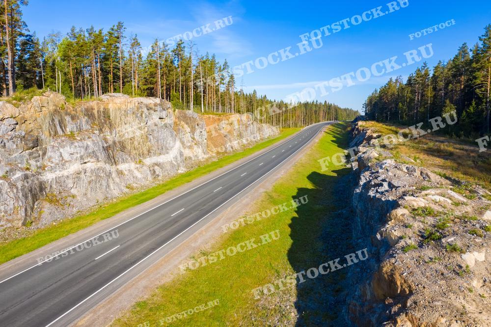 На фотографии изображен участок автодороги в Карелии, проложенной через скалистую местность. С одной стороны дорогу обрамляют отвесные скалы, с другой — сосновый лес. Этот живописный участок дороги символизирует гармонию между природой и инженерным искусством. Карельские дороги, такие как этот участок, обеспечивают удобный доступ к отдаленным уголкам природы и являются важной частью инфраструктуры региона, связывая его с остальной Россией.