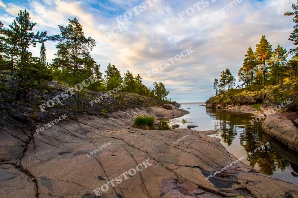 На фотографии представлено Ладожское озеро с его спокойными водами, где камни на берегу и в воде отражаются, создавая уникальный зеркальный эффект. Гладкая поверхность озера словно копирует небесную синь, а камни, слегка погруженные в воду, добавляют гармонии в этот северный пейзаж. Карелия в своем лучшем проявлении — красивое и спокойное место для отдыха на природе.