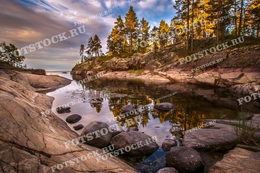 На фотографии изображена тихая бухта Ладожского озера в Карелии, запечатленная в утренние часы. Скалистые берега и густой лес, освещенные первыми лучами солнца, отражаются в спокойной воде, создавая живописный пейзаж. Это место идеально подходит для уединенного отдыха и наслаждения природной красотой. Лесистые вершины и гладкие скалы подчеркивают величие и нетронутость природы Карелии.