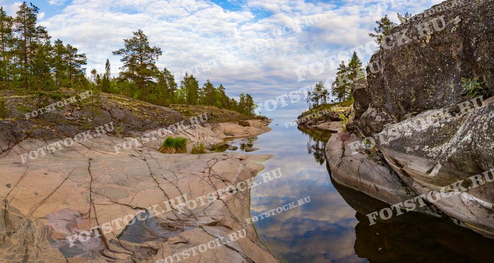 На фотографии изображен скалистый берег Ладожского озера, где среди камней и сосен притаилась тихая заводь. Вода в озере настолько спокойная, что отражает небо и облака, словно зеркало. Такие уединенные места Карелии привлекают любителей природы и фотографов своей уникальной и первозданной красотой. Скалы, вода и леса создают неповторимый ландшафт, характерный для северных регионов России.