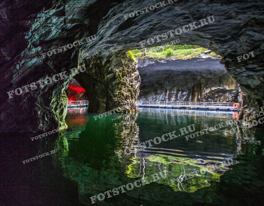 Эта фотография показывает внутреннее пространство пещеры с водной поверхностью, видимое из тёмного входа. Вдалеке освещённый красным светом участок подчёркивает текстуру скальных образований и придает изображению таинственный и мистический настрой. Вода кажется спокойной, а её зелёный оттенок создаёт контраст с тёмными оттенками скал. Фотография идеально подходит для туристических каталогов, исследований геологии, оформления статей о спелеологии или как уникальный элемент декора в помещениях.