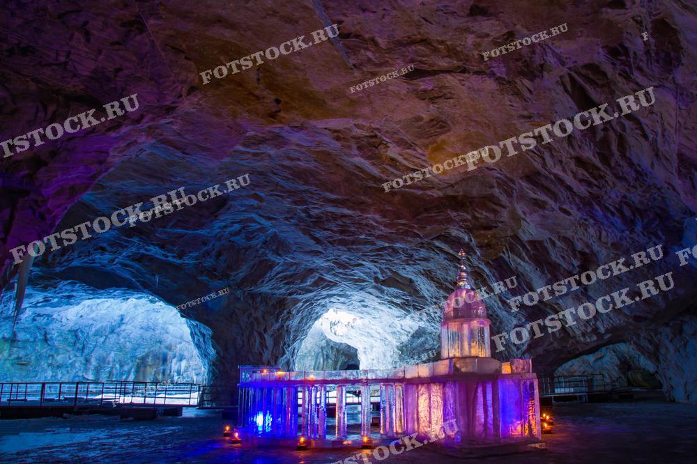 Фотография представляет собой уникальную сцену в парке Рускеала в Карелии — светящийся ледяной дворец внутри исторического мраморного карьера. Подсветка придаёт месту мистическое и волшебное ощущение, а массивные стены пещеры создают впечатляющий фон. Изображение подойдёт для продвижения туристического места, в статьях о путешествиях по России и как визуальный контент для историй о зимних путешествиях.