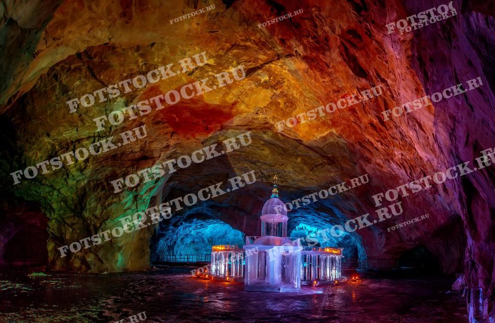 На фотографии изображена пещера в горном парке Рускеала, расположенном в Карелии. В центре кадра видна ледяная часовня, окруженная разноцветными огнями, которые освещают своды пещеры и создают магическую атмосферу. Горный парк Рускеала известен своими мраморными карьерами и подземными пещерами, привлекающими туристов круглый год.