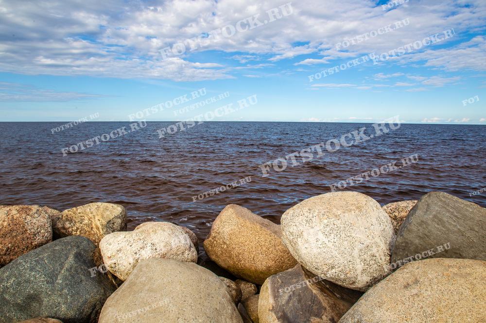 На фотографии изображен каменистый берег Ладожского озера, одного из крупнейших озер Европы. Большие округлые камни служат естественной границей между берегом и водной гладью, создавая впечатление прочности и устойчивости. Ладожское озеро известно своей суровой красотой, и этот пейзаж отражает типичный вид его берегов. Спокойное небо с легкими облаками дополняет ощущение покоя и широты пространства, характерного для Карелии.