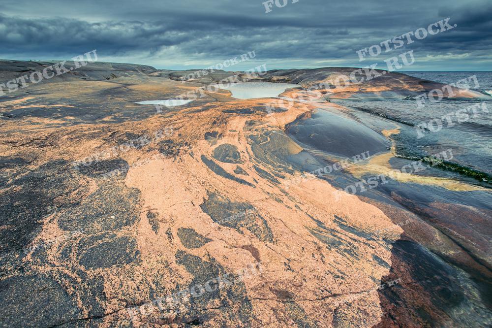 На фотографии изображены скалистые берега Ладожского озера, одного из крупнейших озер в Европе. Уникальные формы и оттенки камней формируют впечатляющий ландшафт, который характерен для карельской природы. Эти берега являются популярным местом для туристов, ищущих уединение и красоту северных пейзажей. Небо, затянутое облаками, подчеркивает дикость и нетронутость этого региона.