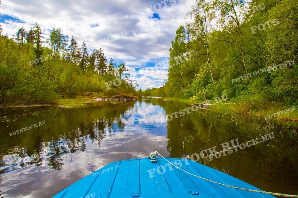 На фотографии изображена речная прогулка на лодке по реке в Карелии. Вид с передней части синей лодки, направленный вдоль тихой реки, окружённой пышной зелёной растительностью и разнообразными деревьями. Небо покрыто облаками, что добавляет изображению насыщенности и глубины. Фотография передаёт ощущение спокойствия и умиротворения, идеально подходит для использования в туристических материалах, статьях о природе и отдыхе на воде.