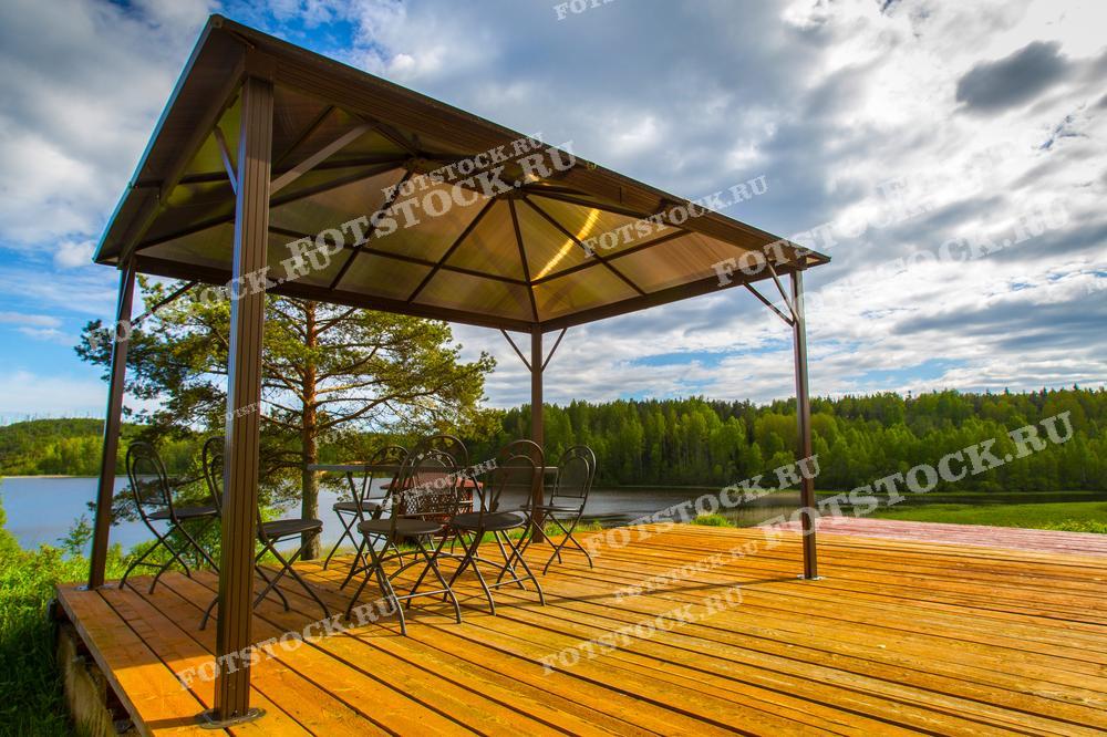 На фотографии изображена беседка, расположенная на деревянной террасе на берегу озера в Карелии. Металлическая конструкция с крышей обеспечивает тень, создавая уютное место для отдыха и общения. За беседкой виден лес и спокойные воды озера, которые придают пейзажу естественную красоту и умиротворение. Это популярное место для летнего отдыха, где можно насладиться природой и свежим воздухом.