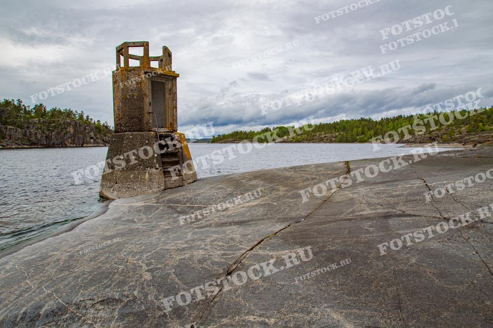 На фотографии изображён старый маяк, стоящий на скалистом берегу Ладожского озера в Карелии. Каменистый берег и спокойные воды под облачным небом создают атмосферу уединения и величия природы. Вдали виднеются лесистые склоны, дополняя картину этого живописного уголка.