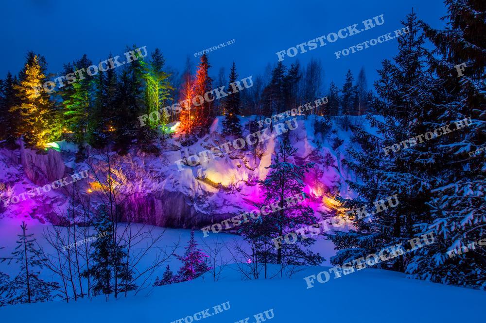 На фотографии изображён горный парк Рускеала зимой, освещённый яркими разноцветными огнями. Заснеженные скалы и деревья выглядят особенно впечатляюще под светом прожекторов, создавая сказочную атмосферу. Это популярное туристическое место в Карелии особенно красиво в зимнее время, когда природа и искусственное освещение соединяются в неповторимое зрелище. Вечерний пейзаж привлекает множество посетителей, желающих насладиться зимней красотой Рускеалы.