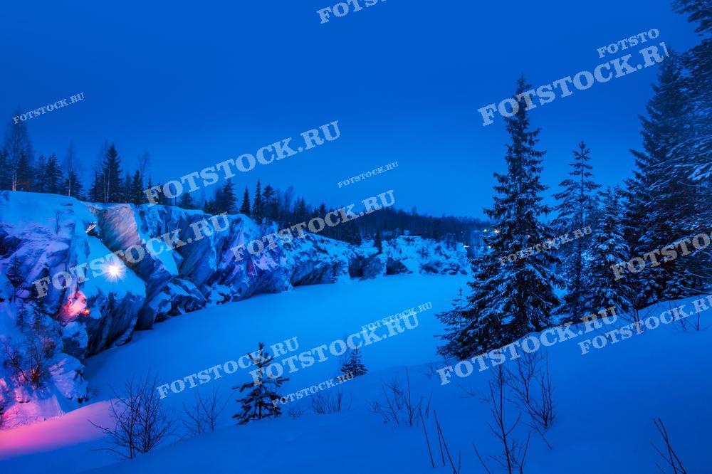 На фотографии изображён ночной зимний пейзаж горного парка Рускеала в Карелии. Заснеженные скалы и деревья освещены мягкой подсветкой, создавая волшебную атмосферу. Снежный покров и тёмное небо подчёркивают красоту этого места.