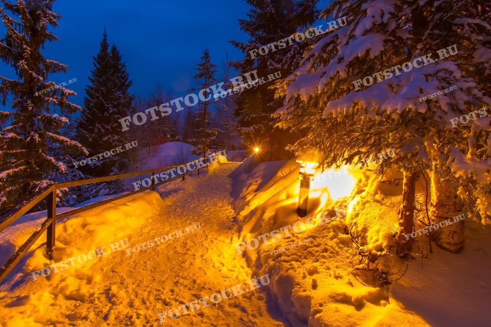 На фотографии изображена зимняя тропа в горном парке Рускеала, освещённая уличными фонарями. Дорога проходит через заснеженный лес, где деревья покрыты толстым слоем снега. Свет фонарей создаёт тёплую и уютную атмосферу, отражаясь на белом снегу и придавая пейзажу волшебный вид. Эта фотография прекрасно иллюстрирует зимнюю красоту Карелии и привлекает туристов своей сказочной обстановкой.