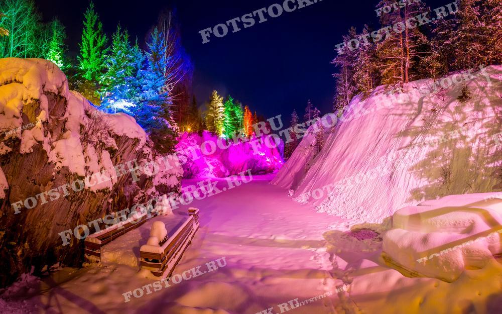 На фотографии изображён горный парк Рускеала зимой, освещённый яркими разноцветными огнями. Заснеженные скалы и деревья выглядят особенно впечатляюще под светом прожекторов, создавая сказочную атмосферу. Ночная подсветка подчёркивает контрасты и текстуры природного ландшафта, превращая его в волшебное зрелище. Это популярное туристическое место в Карелии особенно красиво в зимнее время, когда природа и искусственное освещение создают неповторимую картину.