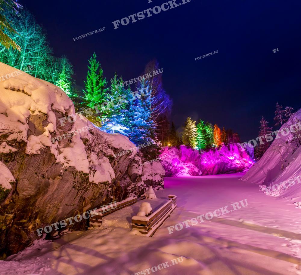 На фотографии изображен зимний пейзаж в горном парке Рускеала, украшенный разноцветной подсветкой, которая освещает скалы и деревья. Снег покрывает все вокруг, усиливая ощущение зимней сказки. Яркие цвета подсветки создают волшебную атмосферу, подчеркивая красоту карельской природы в ночное время. Горный парк Рускеала — одно из самых живописных мест Карелии, особенно в зимний период, когда природа преображается под снежным покровом и яркими огнями.