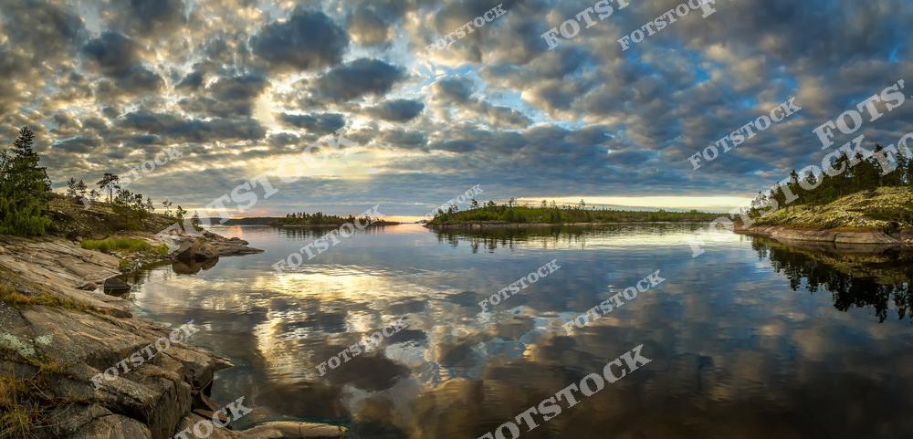 Фотография передаёт атмосферу северного заката над Ладожскими шхерами в Карелии. Разноцветные облака заполняют небо, отражаясь в гладкой водной поверхности, создавая эффект зеркала. Каменистый берег и небольшие острова подчёркивают величие природы Ладожского озера. Это место, где можно почувствовать единение с природой и насладиться её безмятежной красотой.