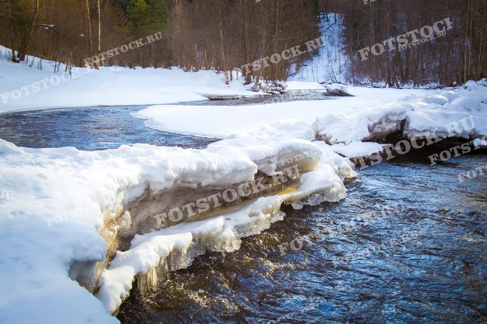 На фотографии изображена зимняя река в Карелии, где вода пробивается сквозь снежный покров, образуя ледяные берега. Снег и лёд окружают русло реки, создавая контраст между тёмной водой и белым ландшафтом. Вдали виднеется лес, добавляя пейзажу глубины и естественной красоты. Этот зимний пейзаж демонстрирует суровость и величие природы Карелии, привлекая внимание своей уникальной атмосферой.