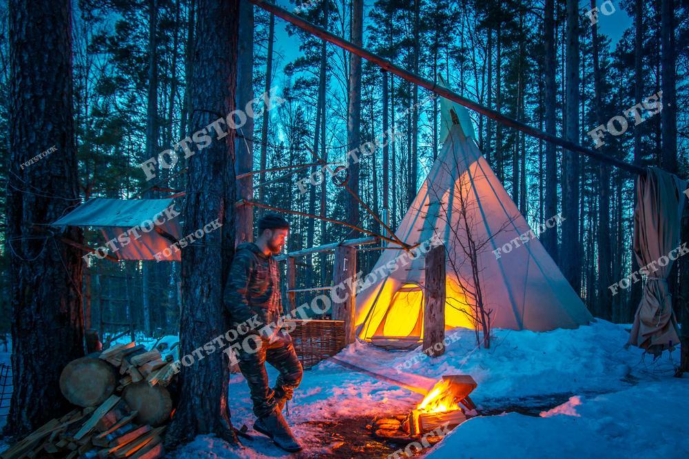 На фотографии изображён туристический этнолагерь в заснеженном лесу Карелии, где установлен вигвам с мягкой жёлтой подсветкой. Мужчина в камуфляже греется у костра на фоне высоких сосен. Возле деревьев сложены дрова, а в воздухе чувствуется тишина и уют зимнего леса. Такие этнопарки и кемпинги особенно популярны в районе Сортавалы и Лахденпохьи в зимний сезон, в том числе в новогодние и рождественские каникулы.
