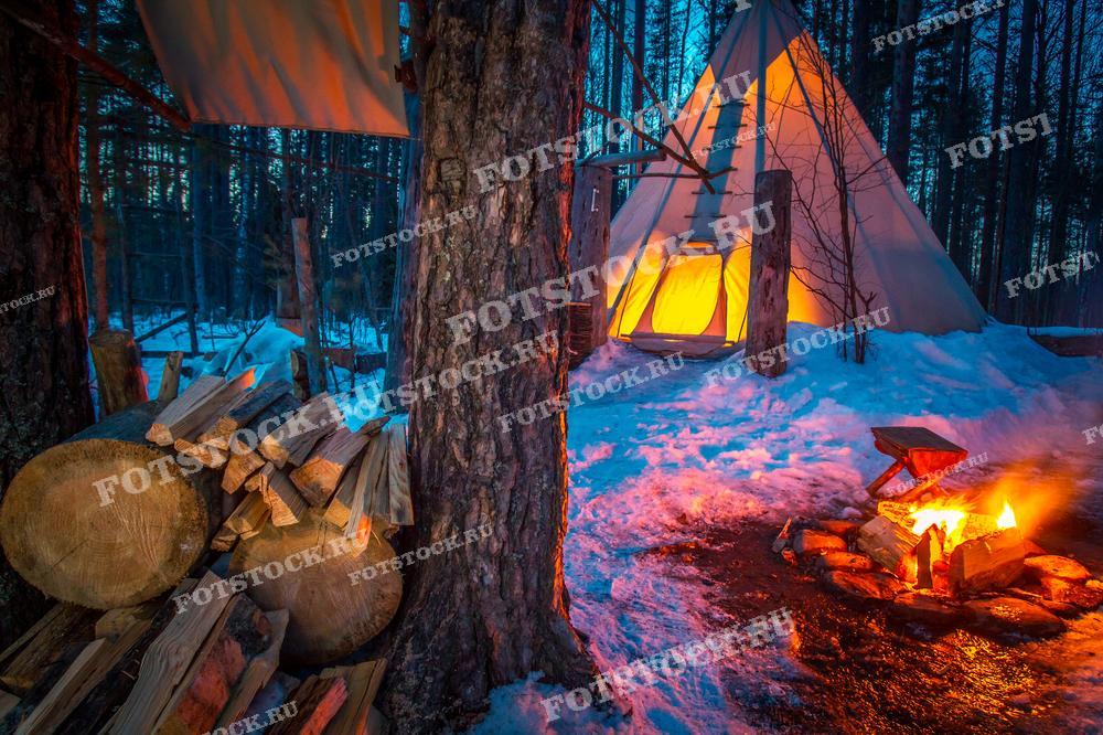 На фотографии изображен зимний лагерь в лесу Карелии. В центре кадра находится палатка, освещенная изнутри теплым светом, рядом горит костер. Дрова аккуратно сложены рядом с деревом, создавая атмосферу уюта и безопасности. Снег на земле подчеркивает зимний сезон и добавляет пейзажу сказочности. Это место идеально подходит для зимнего кемпинга и наслаждения природой Карелии.