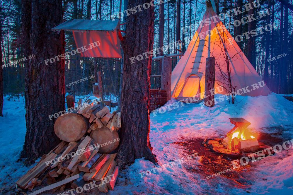 На фотографии изображён зимний лагерь в лесу Карелии, освещённый костром. В центре кадра — типи с тёплым внутренним светом, рядом с которым горит костёр, создавая уютную и тёплую атмосферу.