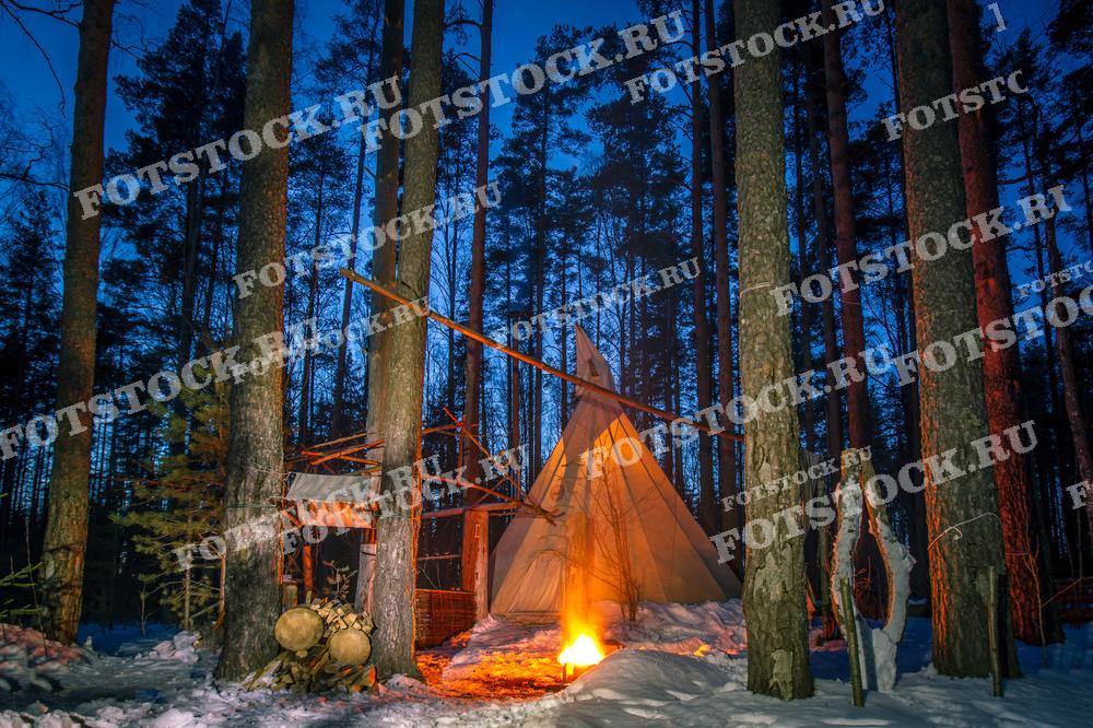 Фотография показывает зимний лагерь в густом карельском лесу. В центре находится палатка в виде традиционного шатра, а перед ней горит костёр, освещая деревья и снег вокруг. Высокие сосны окружают лагерь, создавая ощущение защищённости и уединённости. Это типичный северный пейзаж Карелии зимой, привлекающий любителей активного отдыха и приключений в дикой природе.