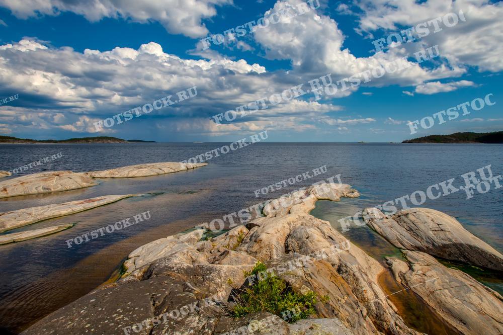 На фотографии изображён живописный пейзаж Ладожского озера в Карелии. Вид открывается на гладкую поверхность воды и изрезанную линию берега, где валуны и скалы покрыты мхом и скудной растительностью. Это изображение может быть использовано в туристических брошюрах, природных журналах или как элемент декора для офисов и жилых помещений, подчеркивая красоту природных ландшафтов России.