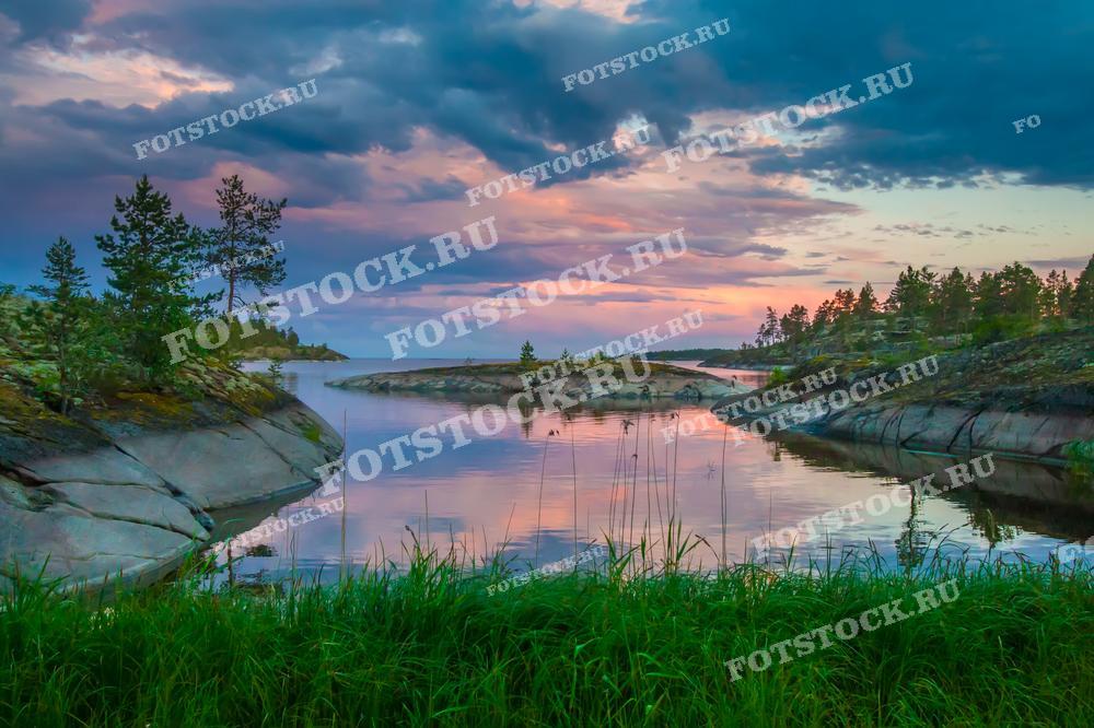 Фотография демонстрирует закат над Ладожским озером в Карелии. Скалистые берега и зелёные деревья обрамляют озеро, создавая красивый и живописный пейзаж. Облака на закатном небе отражаются в спокойной воде озера, добавляя глубины и гармонии в вид. Ладожские шхеры славятся своей природной красотой и привлекают множество туристов.