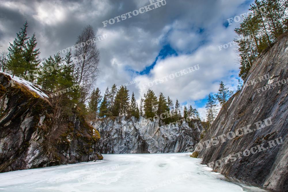 На фотографии изображен замёрзший мраморный карьер в горном парке Рускеала, Карелия. Высокие скалы, окруженные заснеженными деревьями, создают впечатляющий зимний пейзаж. Ледяная поверхность карьера отражает облачное небо, придавая сцене спокойствие и величие. Горный парк Рускеала известен своими мраморными каньонами и является популярным туристическим объектом.