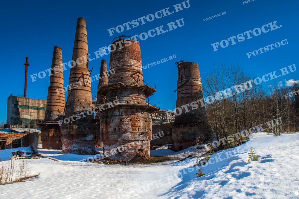 На фотографии изображены старые промышленные печи на итальянском заводе в Карелии. Массивные кирпичные конструкции стоят на заснеженном поле, контрастируя с ярко-синим небом. Этот завод является историческим памятником, свидетельствующим о промышленном прошлом региона.
