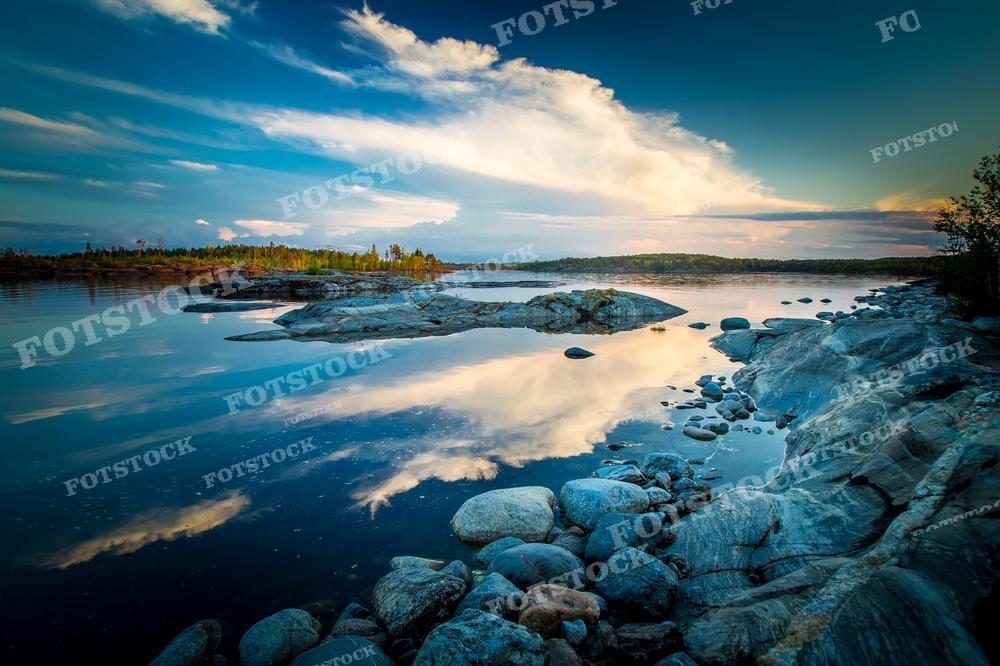 На фотографии представлен захватывающий вечерний пейзаж: спокойная водная гладь озера в Карелии, которая отражает небо с облаками, на горизонте виднеются лесистые острова. На переднем плане видны скалы и камни разного размера. Это изображение подойдет для иллюстрации статей о природе, для создания постеров и открыток, а также может использоваться в качестве фона для экранов и веб-сайтов.